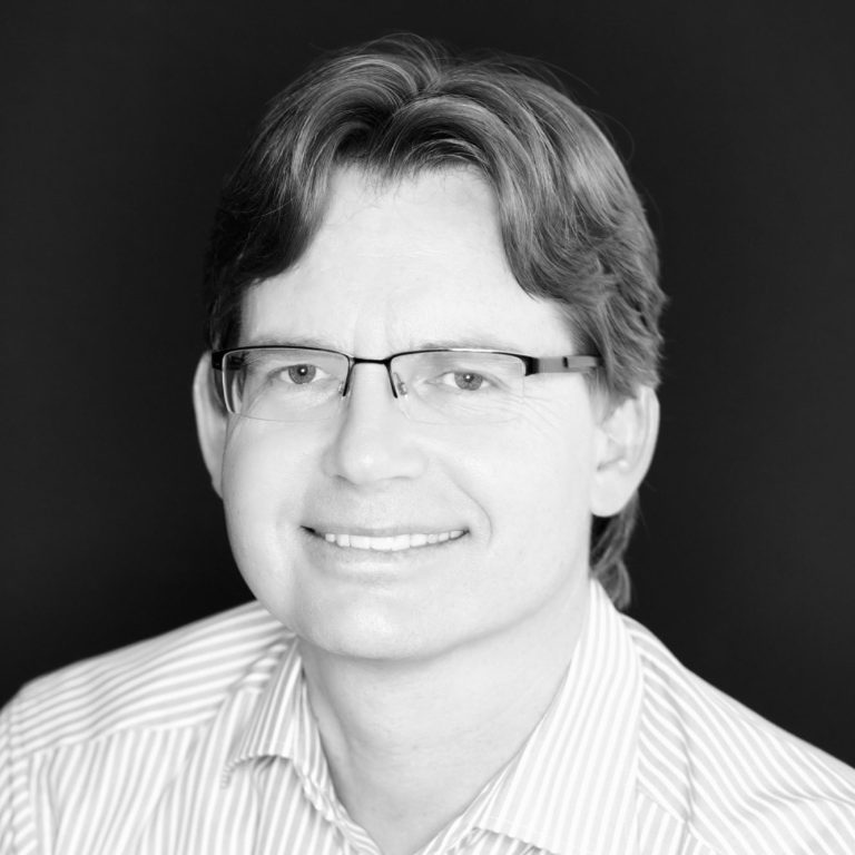 Firmeninhaber_Karl_Kogler Porträtfoto eines lächelnden Mannes mit Brille, gestreiftem Hemd und mittellangem Haar, vor schwarzem Hintergrund.